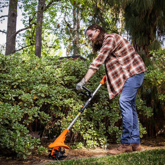 Man using electric rotary tiller in garden with shrubs and trees in background