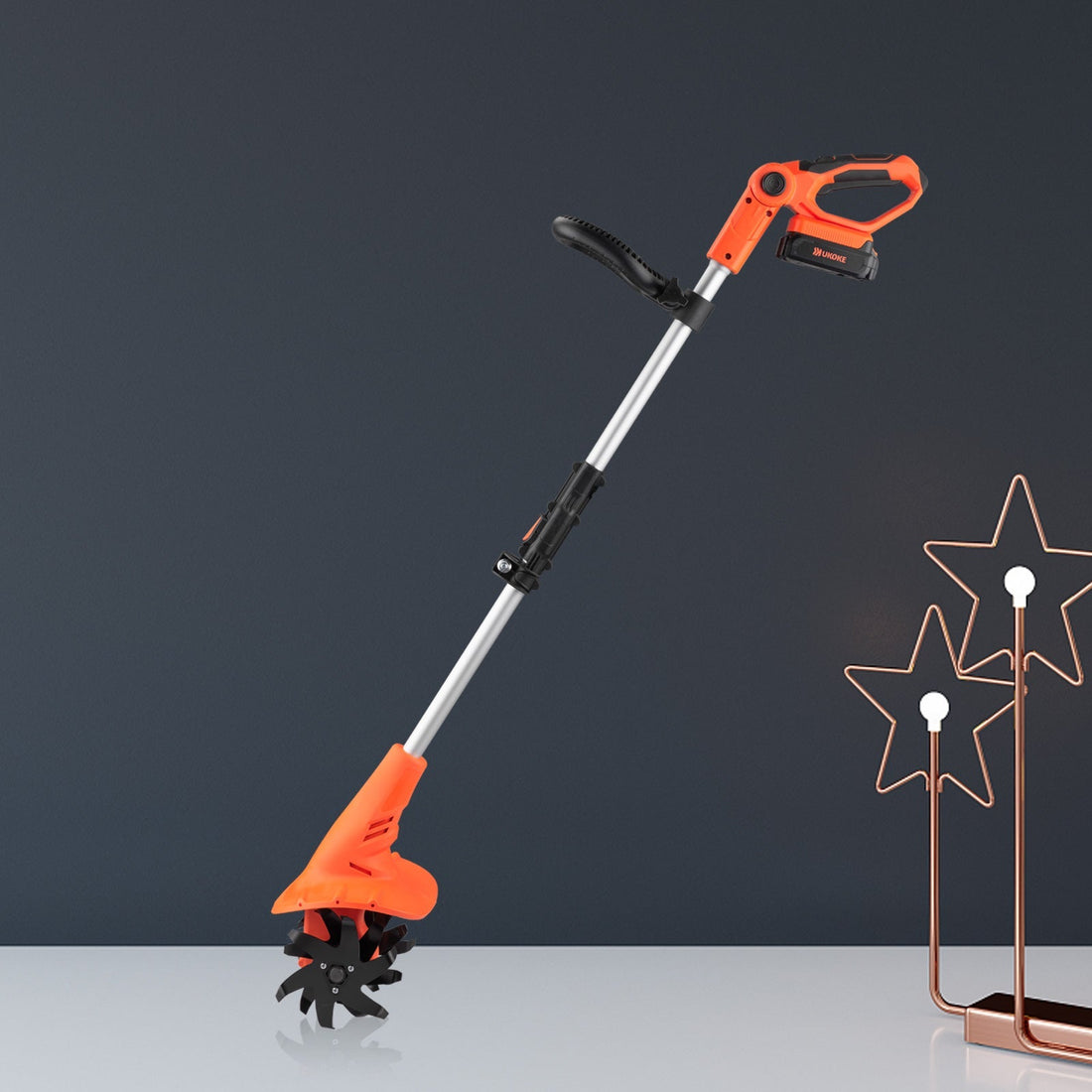 Cordless electric garden tiller with orange handle on a modern table, star lamp decor